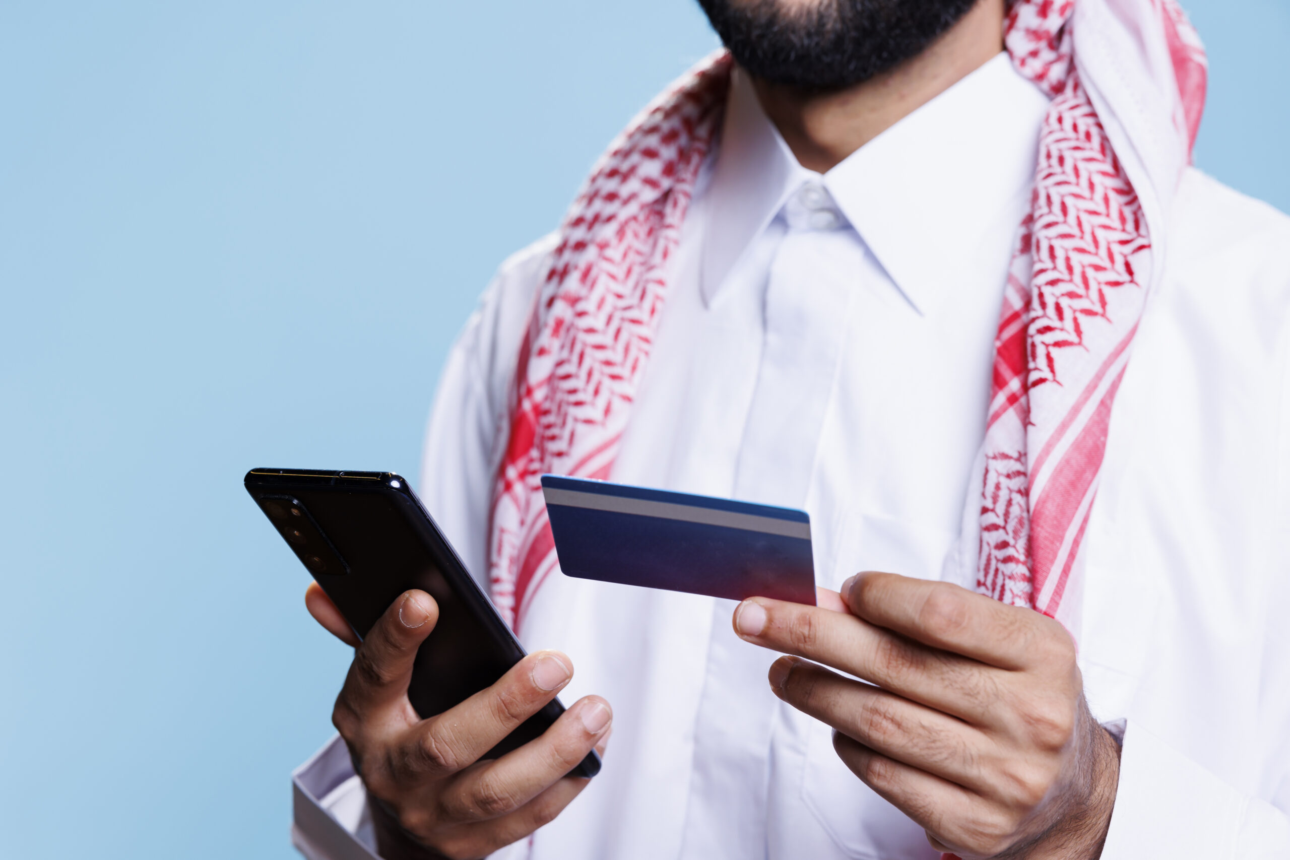 Arab man hands holding credit card and smartphone while making purchase in internet store. Muslim person in traditional clothes shopping online and processing electronic payment closeup